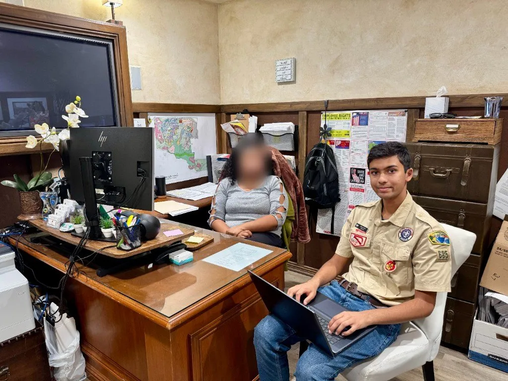 Aditya in his scout uniform working on a laptop at the Del Sur Community HOA office; the other person's face is blurred