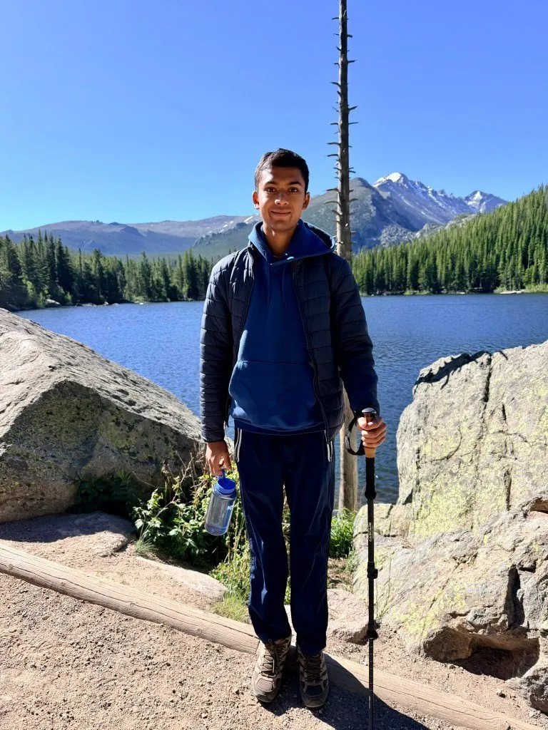 Aditya with a trekking pole in front of an alpine lake and snowy peaks